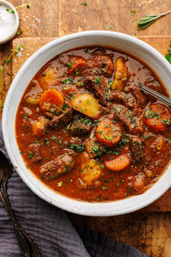 A white bowl of beef stew arranged on a wooden board with a dark napkin and herbs decorated around it.