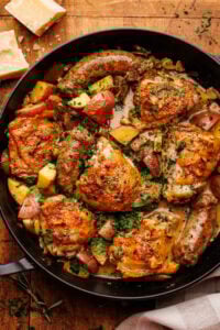 A black cast iron skillet pan with chicken scarpariello with sausage and potatoes in it. The pan is on a wooden background with a block of Parmigiano Reggiano and a light napkin decorated around the pan.