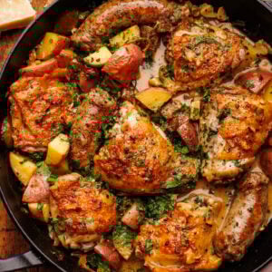 A black cast iron skillet pan with chicken scarpariello with sausage and potatoes in it. The pan is on a wooden background with a block of Parmigiano Reggiano and a light napkin decorated around the pan.
