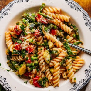 A white dish with pasta primavera and a fork in it arranged on a wooden brown board with a napkin, a glass of white wine, and pansy flowers decorated around the dish.