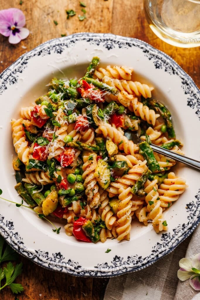 A white dish with pasta primavera and a fork in it arranged on a wooden brown board with a napkin, a glass of white wine, and pansy flowers decorated around the dish.