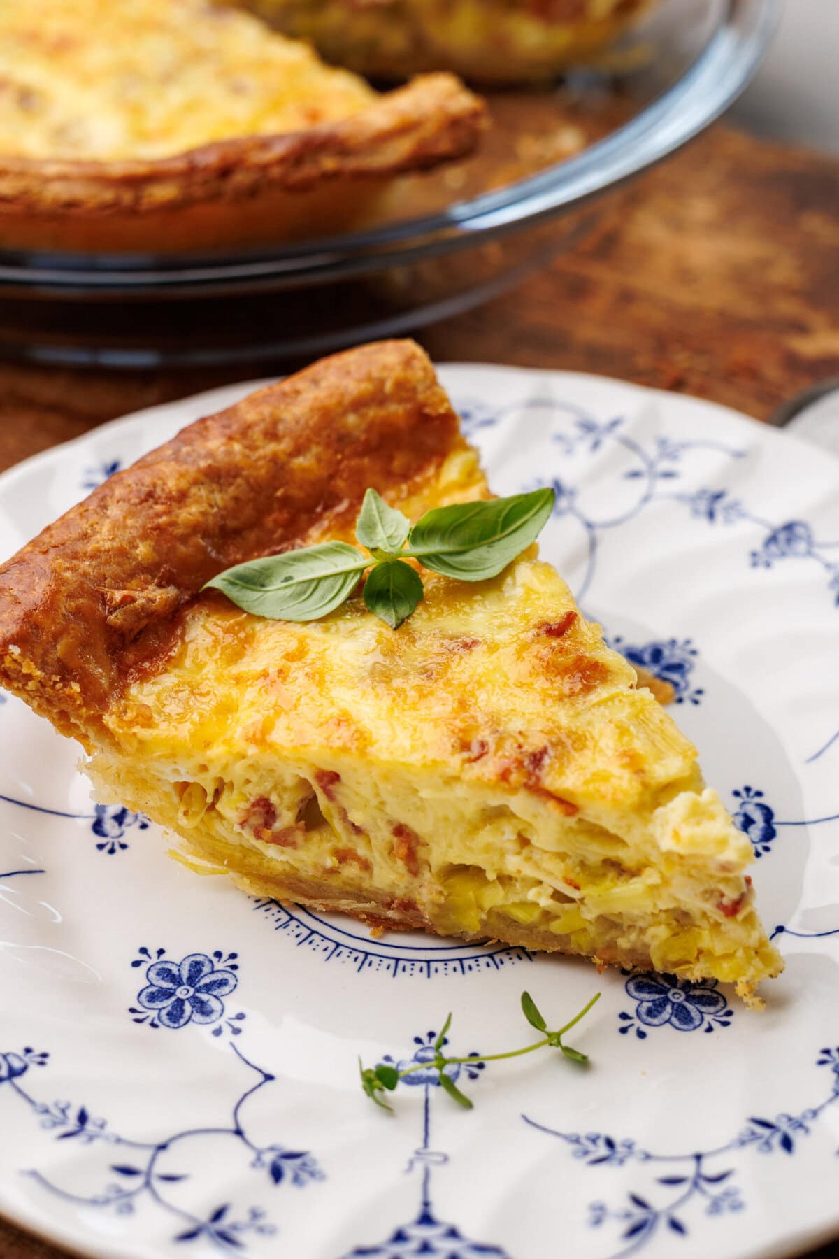 A slice of quiche lorraine on a vintage plate with the rest of the quiche in the background in a glass pie dish