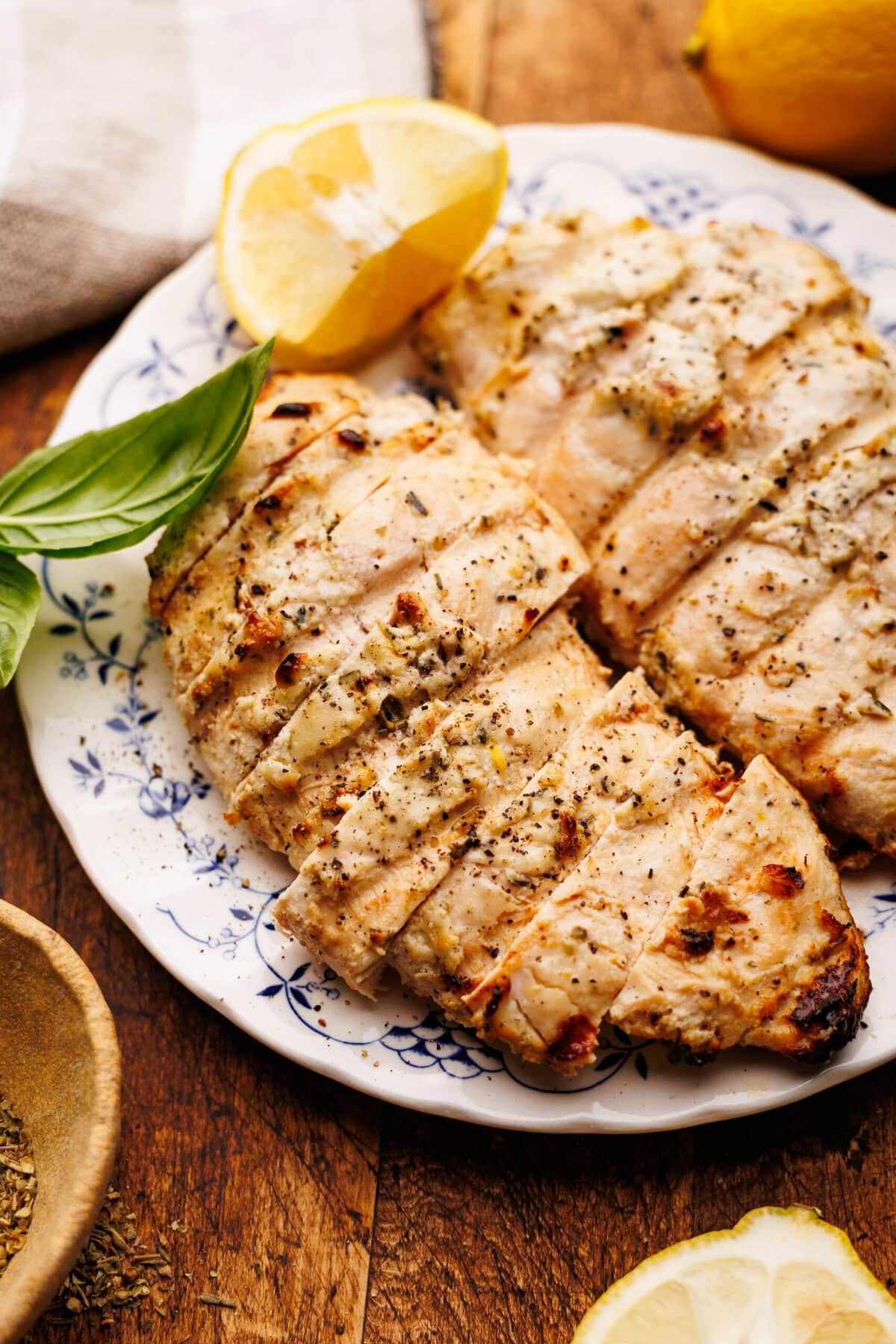 A close up of grilled lemon chicken sliced on a vintage white and blue plate. The plate of chicken is displayed on a brown wooden board with lemons and a napkin placed around it.