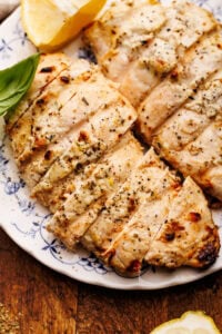 A close up of grilled lemon chicken sliced and displayed on a white and blue vintage plate thats on a wooden board.