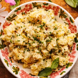 A bowl of potato salad arranged on a wooden board with fresh herbs, flowers, and a grey gingham napkin arranged around it.