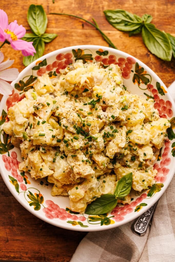 A bowl of potato salad arranged on a wooden board with fresh herbs, flowers, and a grey gingham napkin arranged around it.
