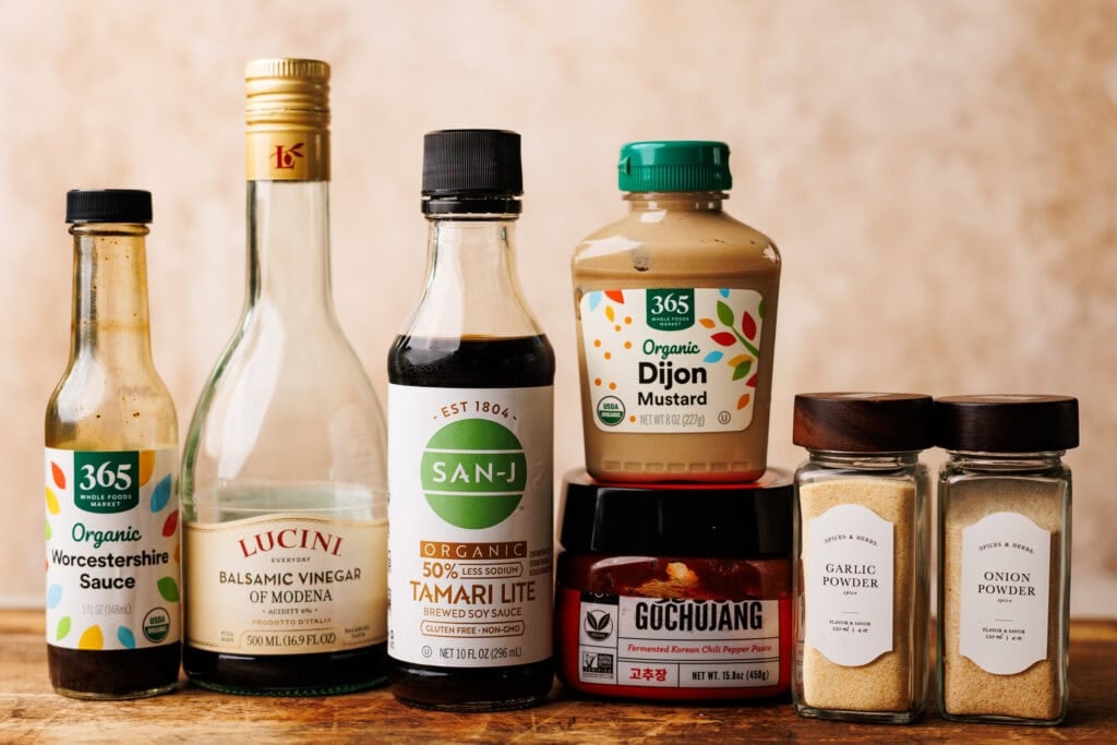 Ingredients and condiments for steak kabob marinade arranged on a wooden table with a tan background behind them.