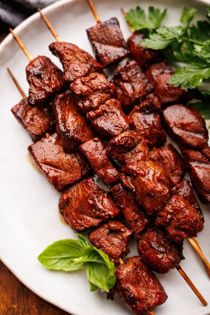 Steak kabob's on a white plate with fresh green herbs placed around them.
