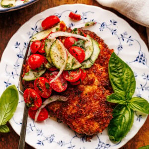 A white vintage plate with chicken cutlets and garden salad on it with fresh basil, and a gingham napkin placed around it. All of this is displayed on a brown wooden board.