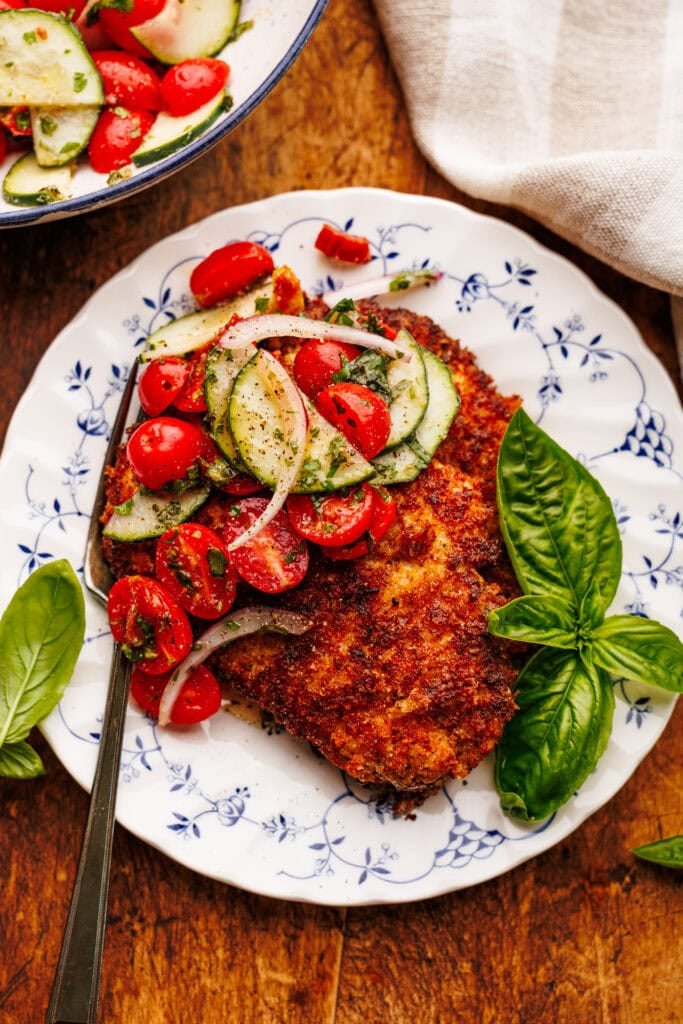 A white vintage plate with chicken cutlets and garden salad on it with fresh basil, and a gingham napkin placed around it. All of this is displayed on a brown wooden board.