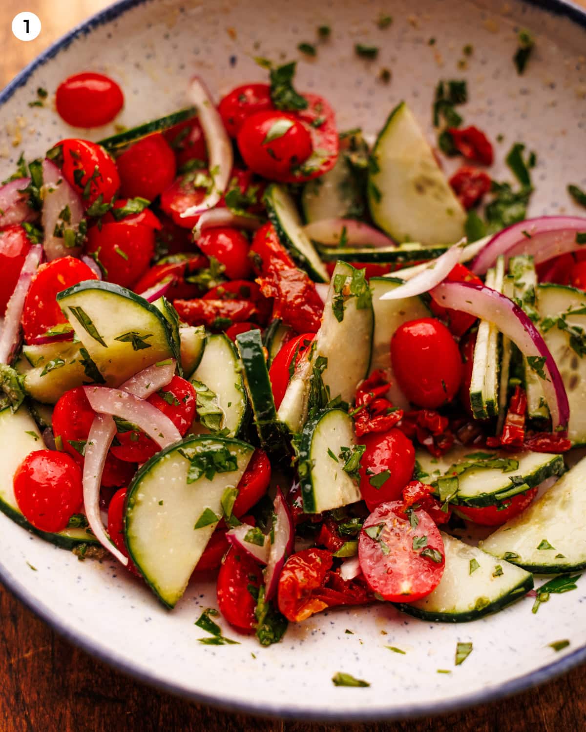 A photo labeled step one showing a garden salad in a white speckled bowl on a wooden surface. The salad has halved cherry tomatoes, sliced cucumbers, red onion, sun-dried tomatoes, and chopped fresh herbs tossed in dressing.