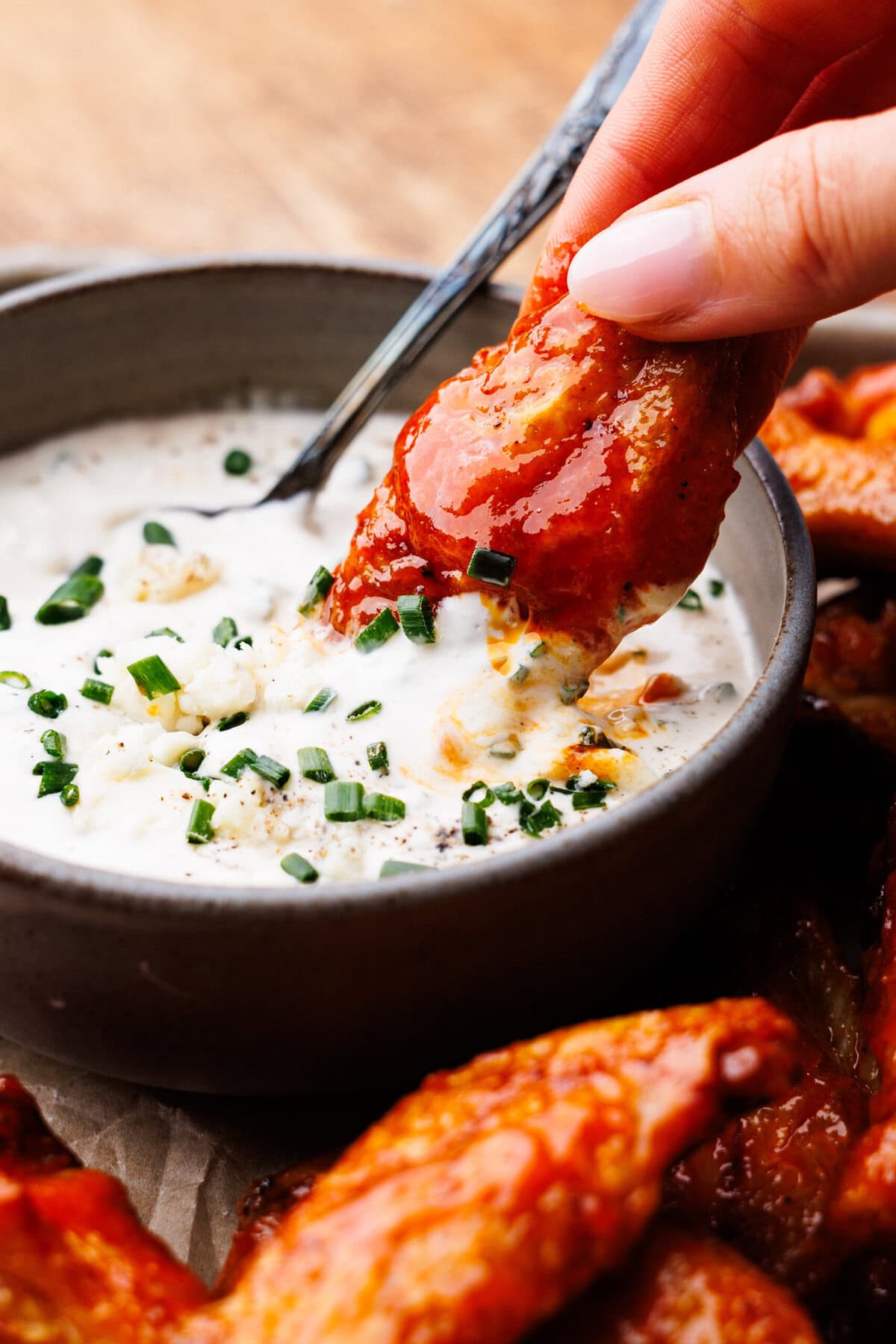A buffalo chicken wing being dipped in a blue cheese sauce in a gray bowl, with more wings below on a wooden cutting board