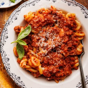 A white bowl with pasta and Italian ragu with a fork in it. The bowl of pasta is displayed on a wooden background with Parmigiano Reggiano, basil leaves, a glass of red wine, and a grey napkin placed around it.