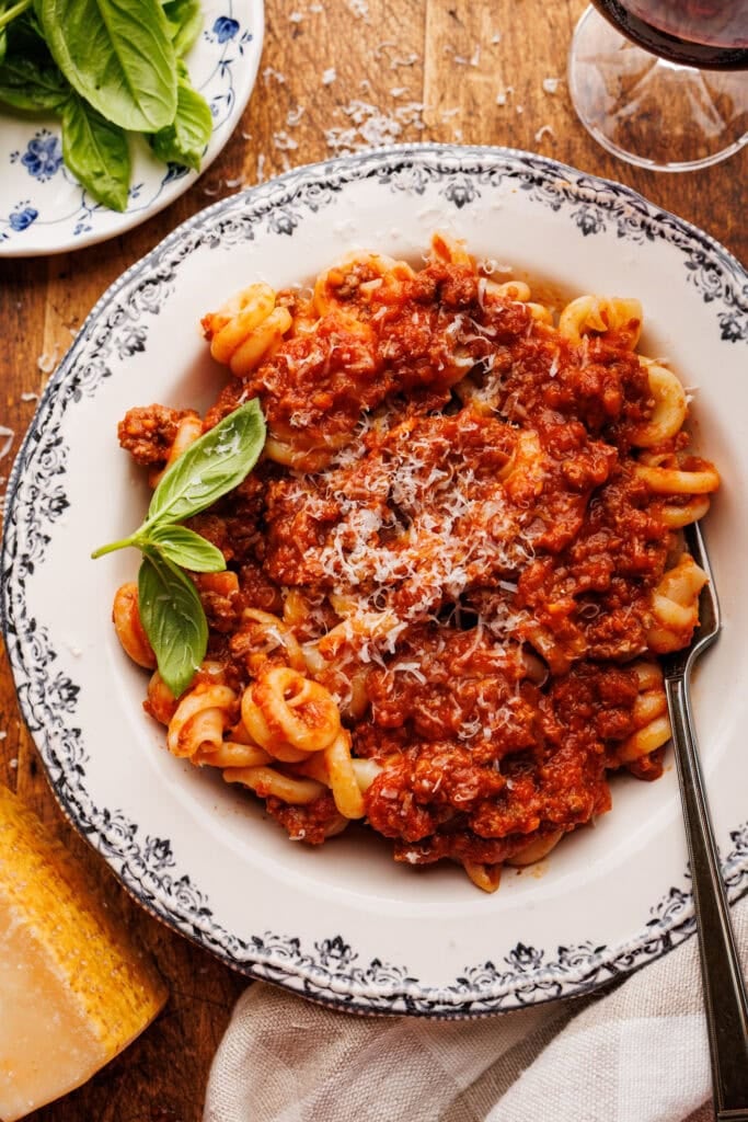A white bowl with pasta and Italian ragu with a fork in it. The bowl of pasta is displayed on a wooden background with Parmigiano Reggiano, basil leaves, a glass of red wine, and a grey napkin placed around it.