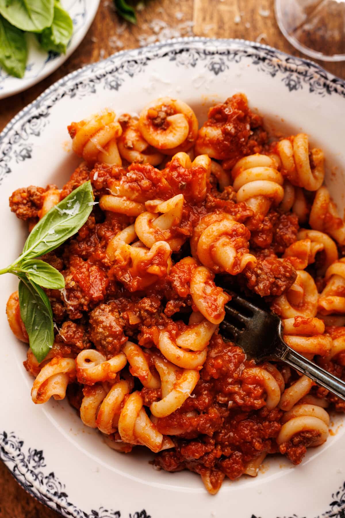 A close up of nodi morini pasta in a bowl with Italian ragu displayed on a wooden board with a glass of wine and plate of fresh basil around it.
